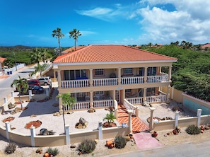A two-story building with a red-tiled roof, multiple balconies, and a landscaped front yard with rocks and plants. - Aruba Oasis with pool, walking distance to the beach, hotels and restaurants (Palm Beach)