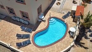 En swimmingpool med solstolar och ett litet bord, omgiven av en innergård med tegelbeläggning och en byggnad med flera fönster.