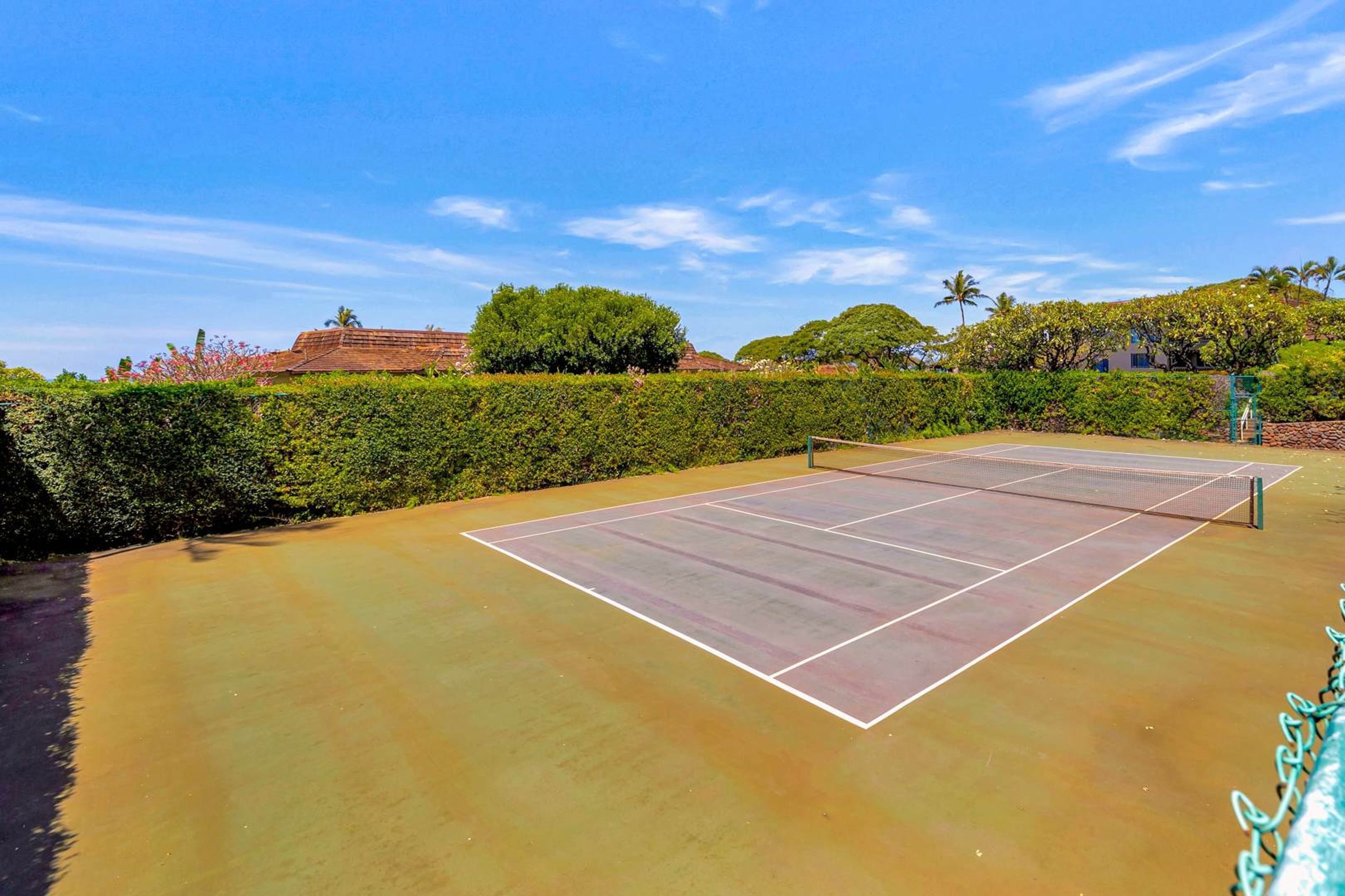 An outdoor tennis court with a net, surrounded by greenery and a clear sky.