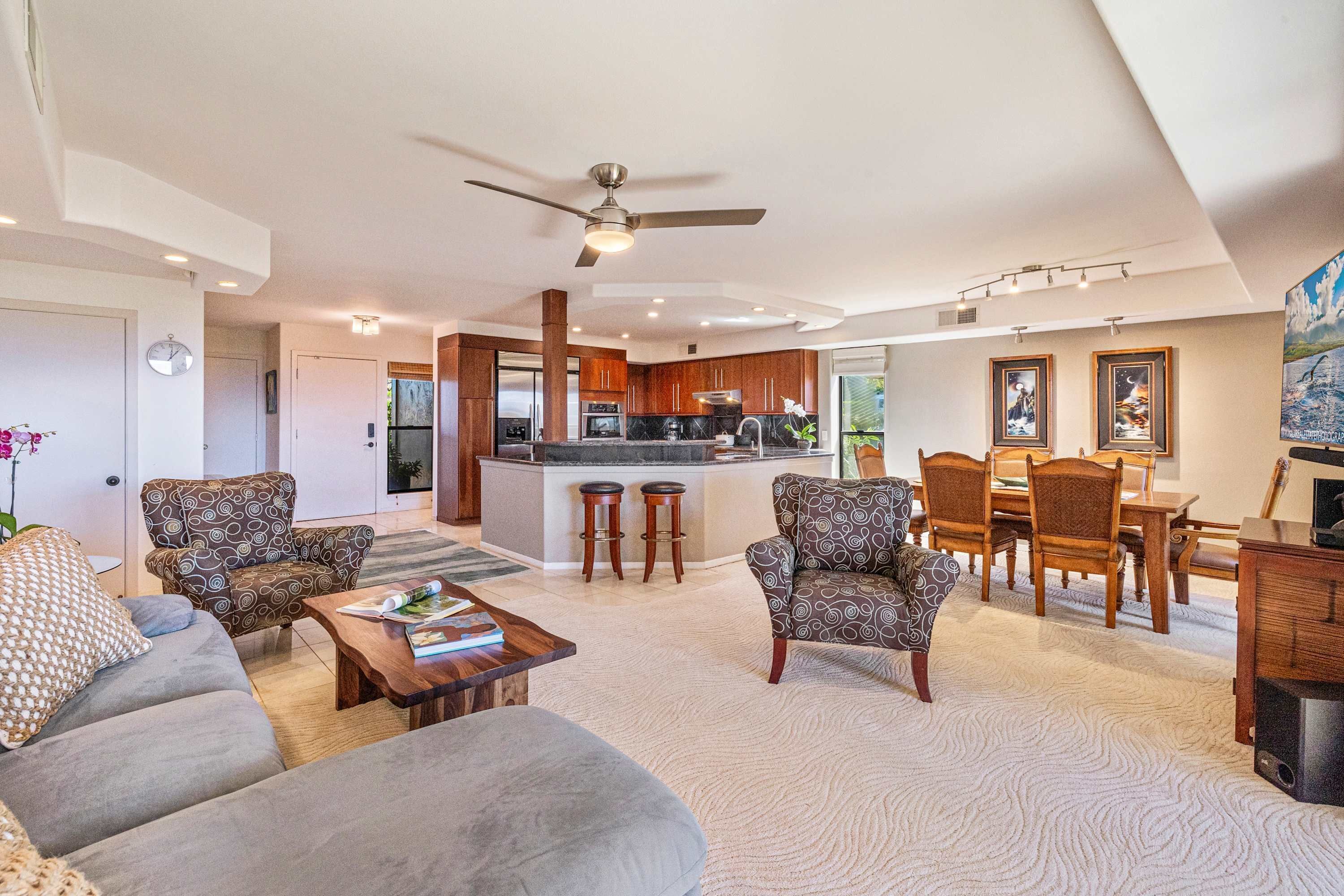 A spacious living room with a grey sofa, patterned armchairs, a wooden coffee table, and a dining area with a wooden table and chairs.