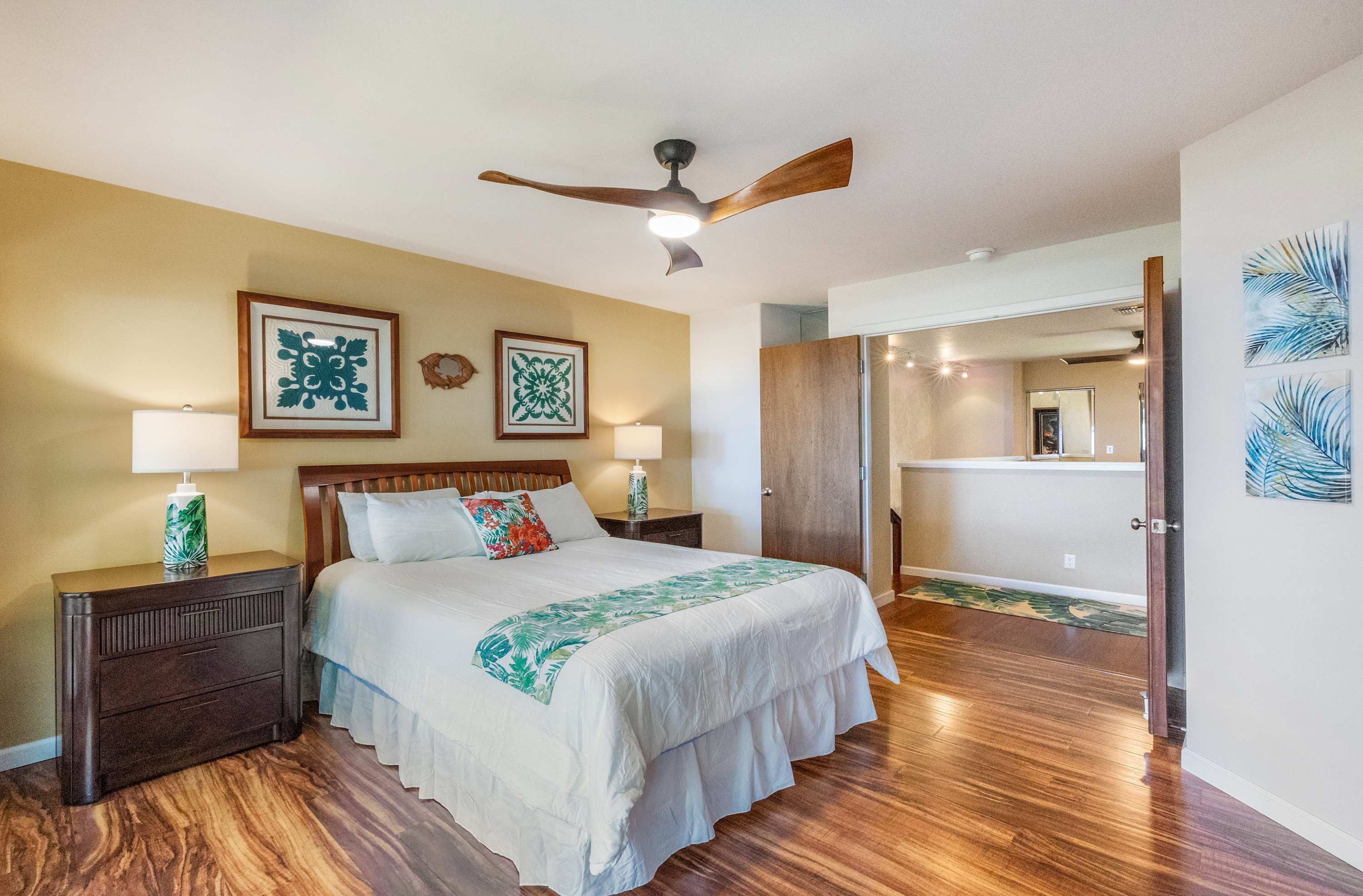 A bedroom with a wooden floor, a bed with a white bedspread, two bedside tables with lamps, a ceiling fan, and artwork on the walls.