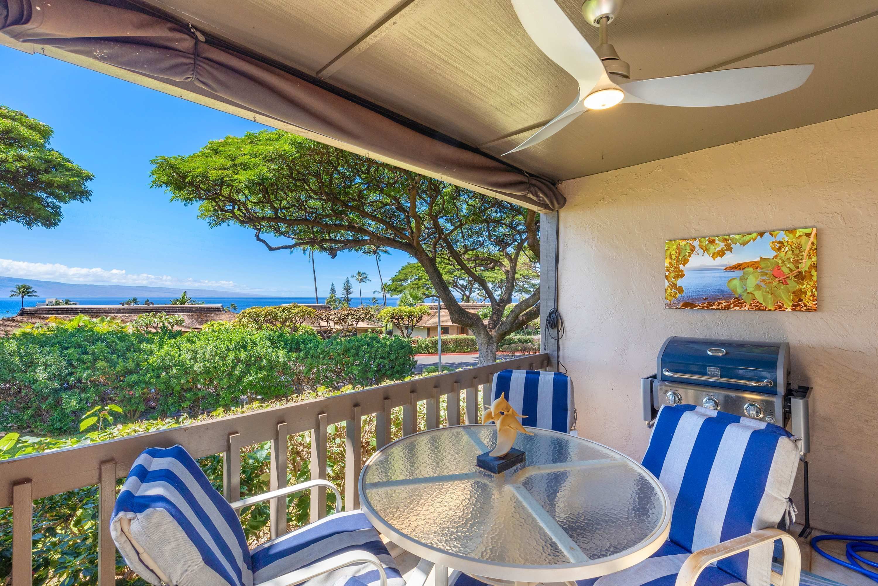 A covered outdoor seating area with a table and chairs, a grill, and a view of the ocean and greenery.