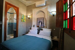 A hotel room with a bed, a shower, a wall-mounted air conditioner, and a window with colored glass panels. - Riad Mazal (Marrakech)