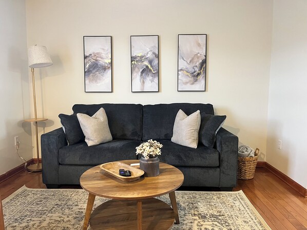 A living room with a dark grey sofa, three abstract paintings on the wall, a floor lamp, a round wooden coffee table with a vase of flowers, and a woven basket. - NEW! 2-bedroom townhome- 0.3 miles from I-81 (Glade Spring)