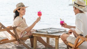 Two people sitting on beach chairs at a wooden table with drinks, overlooking a calm sea with boats and a mountain in the background. - Careyes (Costa Careyes)
