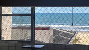 A view through a window with a chain-link fence, overlooking a beach with waves and a distant ship. - Vacation/Carnival - SEA VIEW 3 Bedrooms - Sleeps 10! (Praia Grande)