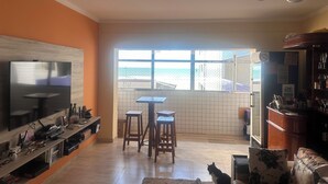 A living room with a TV, wooden shelves, a dining area with stools, and a view of the sea through a large window. - Vacation/Carnival - SEA VIEW 3 Bedrooms - Sleeps 10! (Praia Grande)