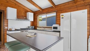 A modern kitchen with wooden paneling, white cabinetry, a large window, and a built-in microwave. - Neptune's Nook - Cape Paterson (Cape Paterson)