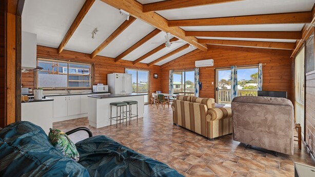 A spacious living area with a kitchen, wooden beams, a sofa, and a dining table. - Neptune's Nook - Cape Paterson (Cape Paterson)