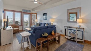 Ein geräumiges Wohnzimmer mit einem blauen Sofa, einem Holztisch und einem kleinen Beistelltisch mit einer Lampe. Es gibt einen Deckenventilator, ein großes Fenster mit Jalousien und eine Tür, die auf einen Balkon führt.