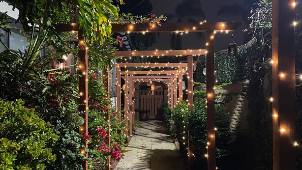 A pathway with a wooden pergola adorned with string lights, surrounded by lush greenery and flowering plants. - Upscale San Diego Home with Secret Room & Private Yard! (San Diego)