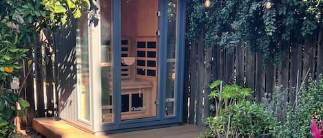 A small wooden structure with glass doors, a wooden deck, and a potted plant in a garden setting.