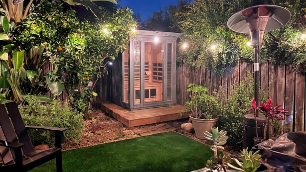 Una pequeña estructura de madera con puertas de vidrio, rodeada por un jardín con diversas plantas y césped.