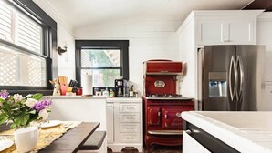 Una cocina con armarios blancos, una estufa roja, un refrigerador de acero inoxidable y una ventana con persianas.