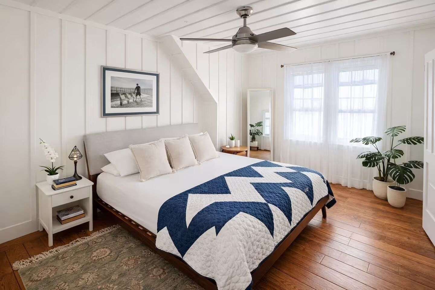 Ein Schlafzimmer mit Bett, Nachttisch, Deckenventilator, einem Fenster mit Vorhängen und einer Zimmerpflanze.