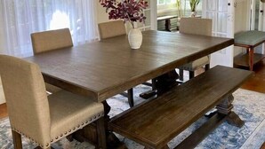 Un comedor con una gran mesa de madera, sillas acolchadas, un banco y un jarrón decorativo con flores.