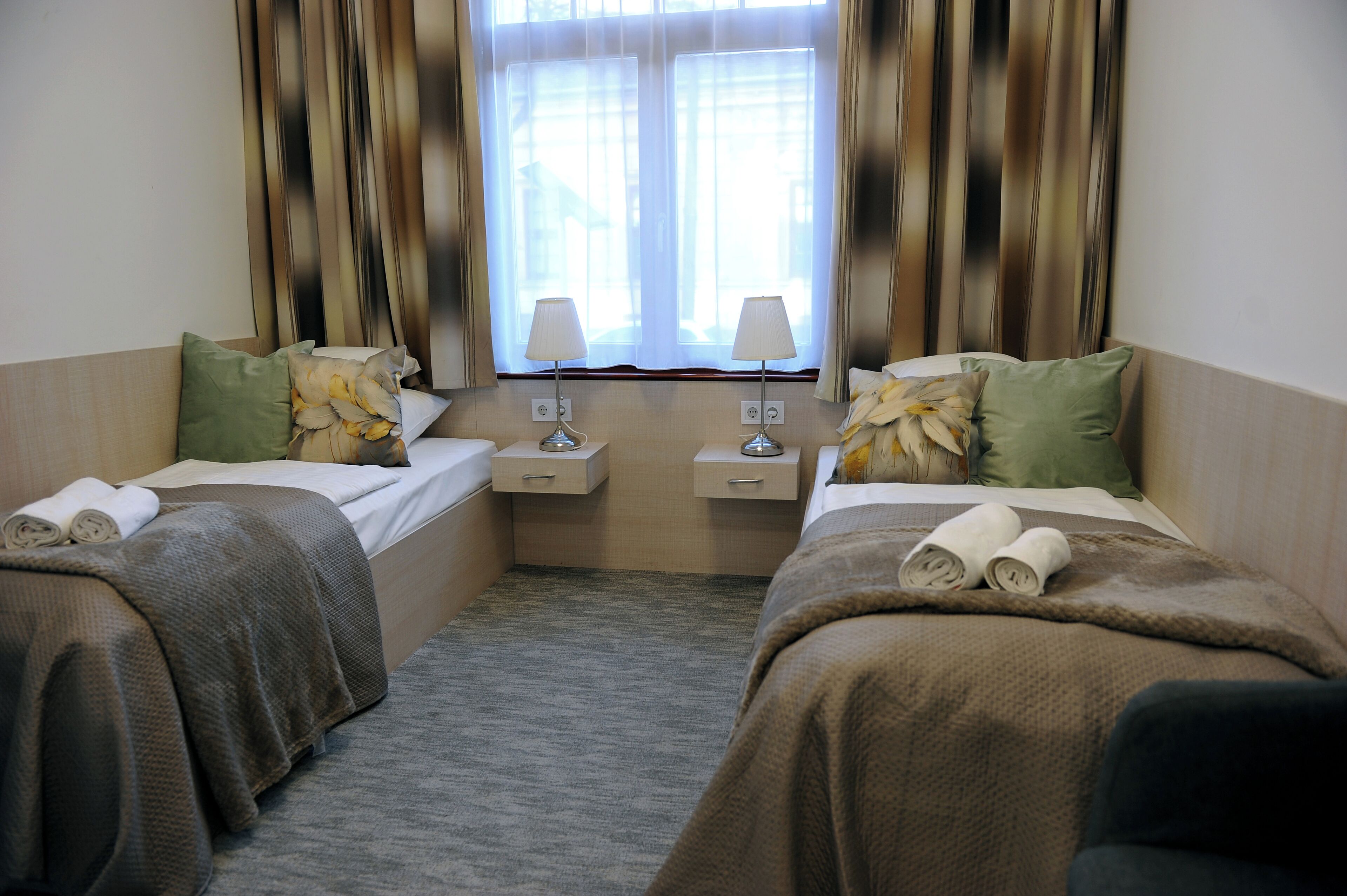 A hotel room with two beds, each with a pillow and a rolled towel. There are bedside tables with lamps and a window with curtains.