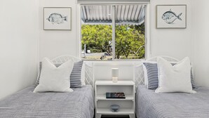 Quarto com duas camas, cada uma com roupa de cama listrada e almofadas brancas. Há quadros com arte de peixes nas paredes, uma janela com vista para o verde e uma mesa pequena com uma lâmpada e um livro.