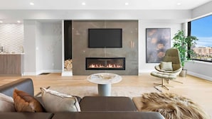 A modern living room with a fireplace, a flat-screen TV, and a large window offering a city view. - Hudson Views (North Bergen)