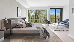A modern bedroom with a large bed, a desk, and a chair. There is a large window with a view of trees. - Hudson Views (North Bergen)
