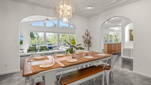 A dining area with a wooden table set for four, white chairs, and a chandelier. - Sleeps 24 | Hot Tub | Firepit | 17 beds | 2 Houses (Oakhurst)