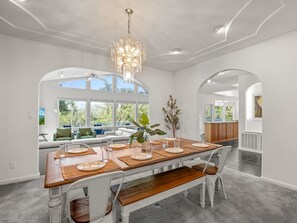 A dining area with a wooden table set for four, white chairs, and a chandelier. - Sleeps 24 | Hot Tub | Firepit | 17 beds | 2 Houses (Oakhurst)