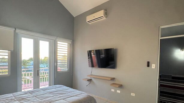 A bedroom with a bed, a ceiling fan, an air conditioning unit, a TV mounted on the wall, and a view of the outside through a window. - Modern Townhouse Near Beaches & BQN Airport with Pool (Aguadilla)