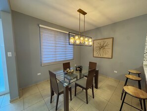 Un coin repas comprenant une table en verre, six chaises, un lustre, une grande fenêtre avec stores et une œuvre d’art abstraite accrochée au mur.
