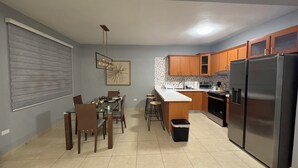 A modern kitchen with wooden cabinets, a stainless steel refrigerator, and a dining area with a glass table and chairs. - Modern Townhouse Near Beaches & BQN Airport with Pool (Aguadilla)