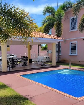 A swimming pool with a tiled deck, a covered seating area with tables and chairs, and palm trees. - Modern Townhouse Near Beaches & BQN Airport with Pool (Aguadilla)