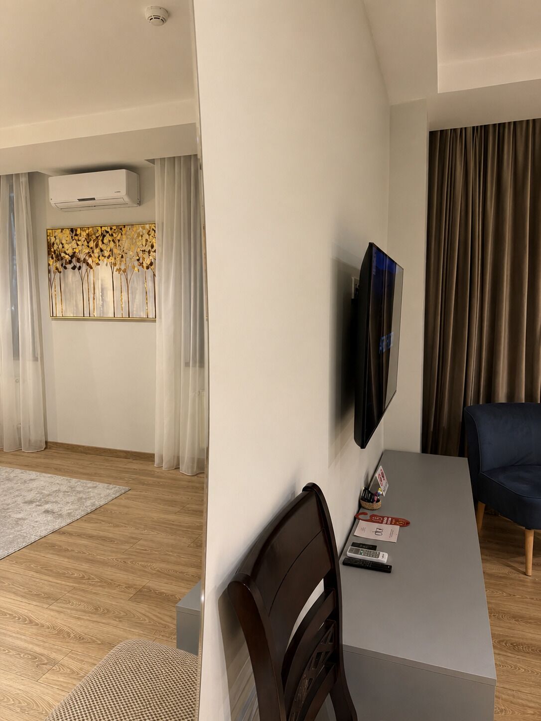 Una habitación de hotel moderna con una televisión de pantalla plana montada en la pared, una mesa de comedor con sillas, una obra de arte decorativa y un gran ventanal con cortinas.