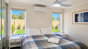A bedroom with a bed, a ceiling fan, an air conditioning unit, a window with a view of a backyard, and a framed picture on the wall.