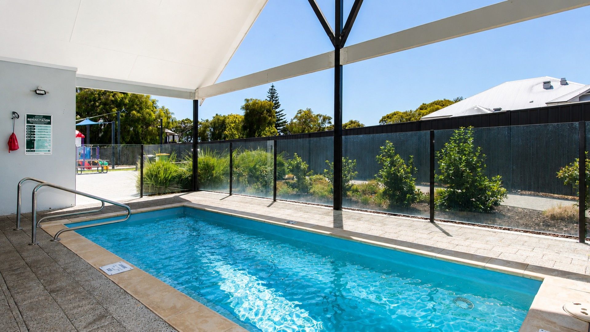 An indoor swimming pool with a clear view of the surrounding area, including a fenced garden and a building in the background.