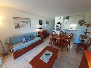 A living room with a sofa, a dining table with chairs, a kitchen area with appliances, and a wall clock. - Very nice 3-room apartment with indoor pool and golf course view in Cap d'Agde (LE CAP D AGDE)