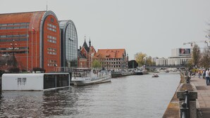 A river with a boat docked on the left, a bridge in the background, and buildings along the riverbank. - Camelia Apartament Balkon Gara (Bydgoszcz)