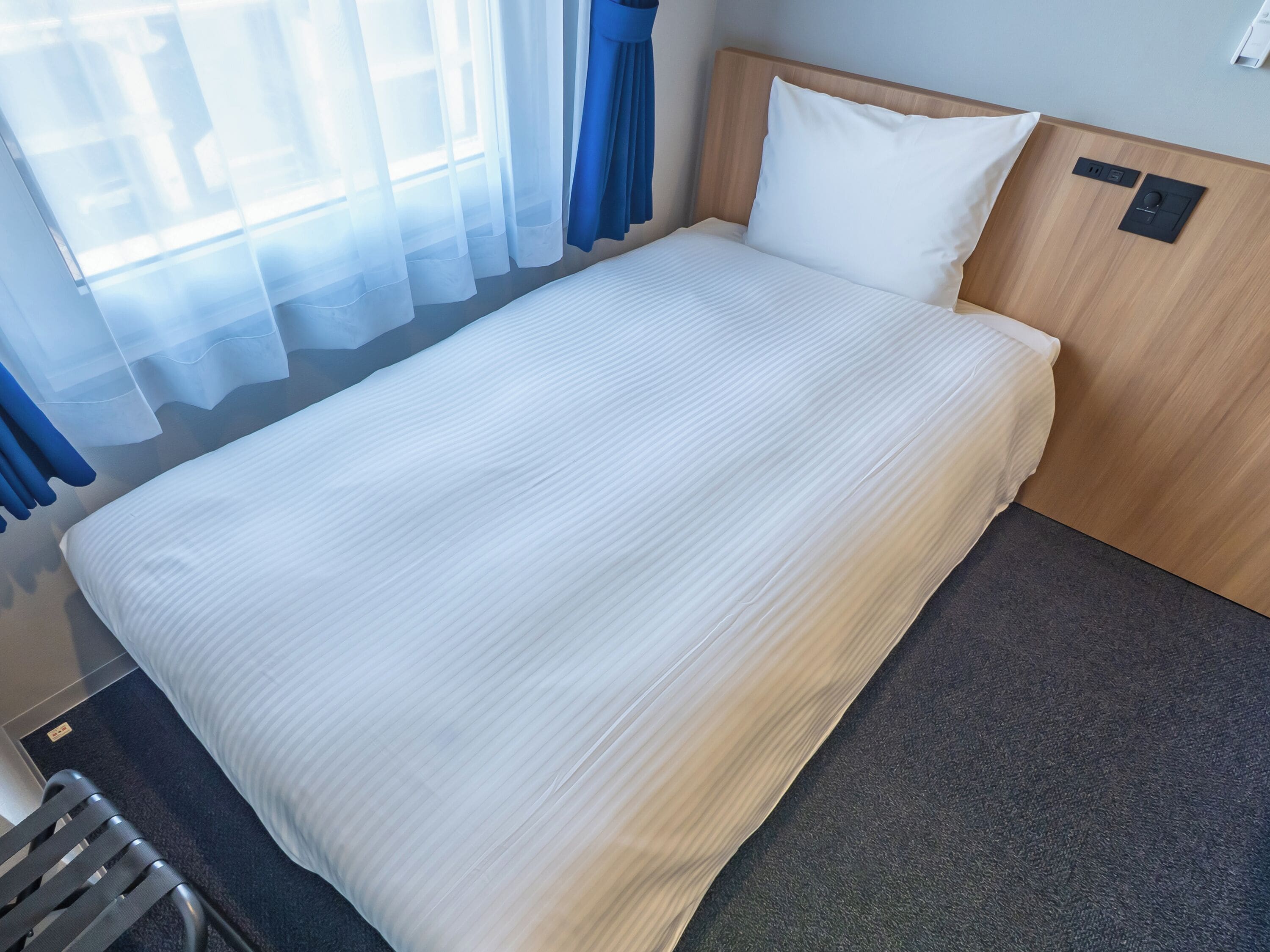 A neatly made bed with a headboard, a bedside table, and a window with sheer curtains.