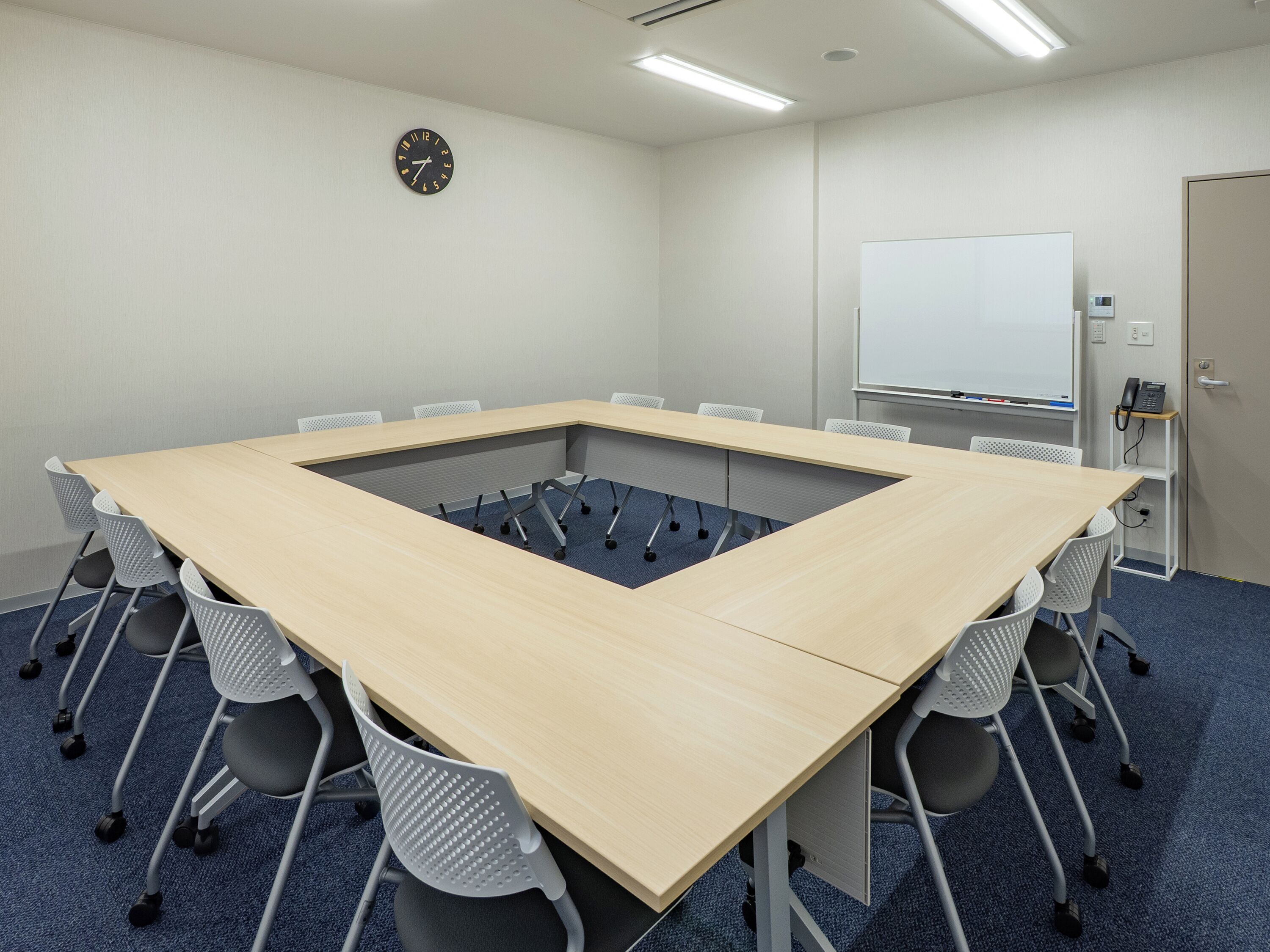 A conference room with a long table, chairs, a clock, a whiteboard, and a phone on a shelf.