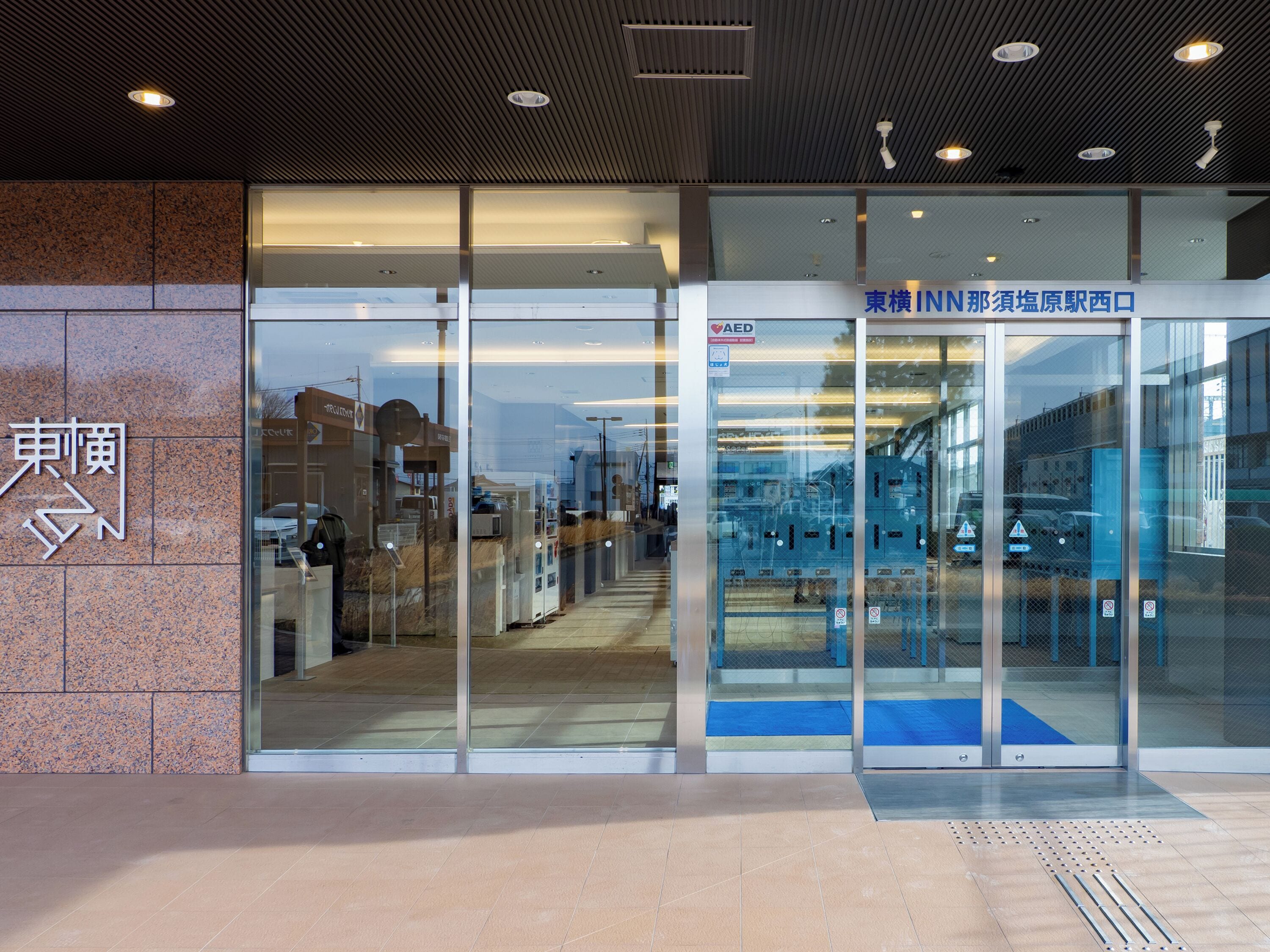 A building entrance with glass doors, a sign with Japanese characters, and a blue carpet leading inside.