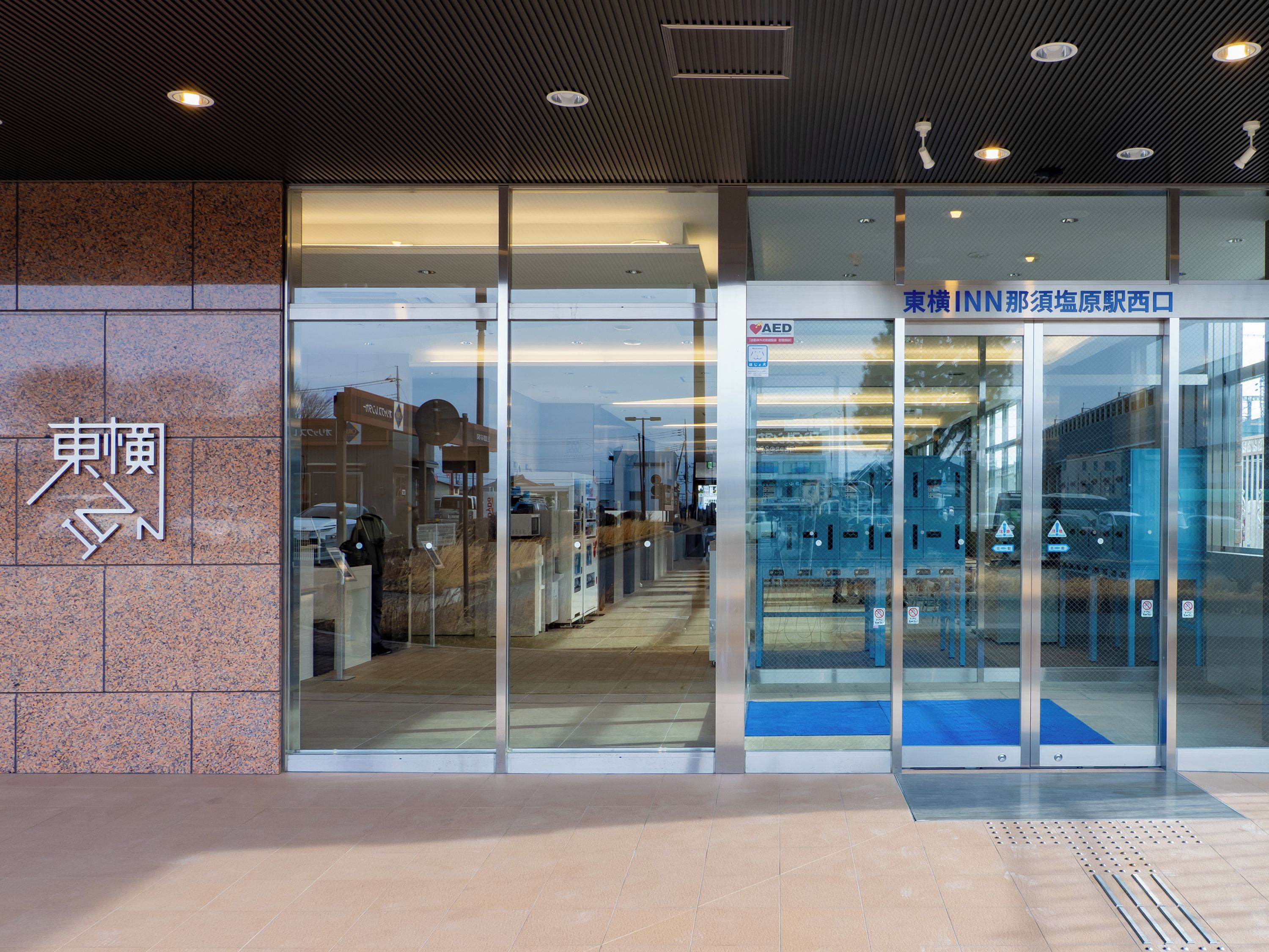 A building entrance with a glass door, a sign with Japanese characters, and a blue mat at the entrance.