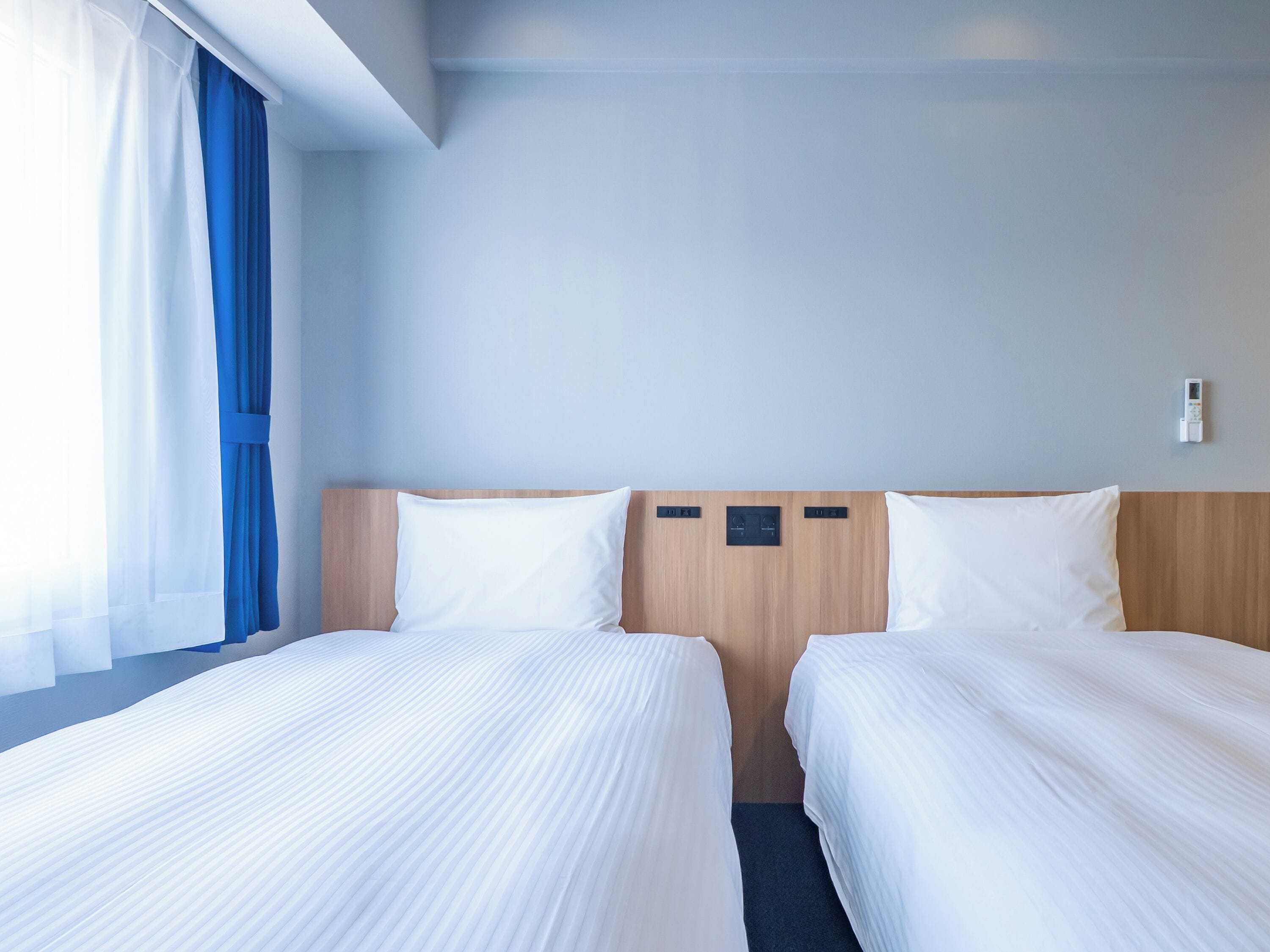 Quarto de hotel com duas camas, cabeceira de madeira, painel de controle e janela com cortinas.
