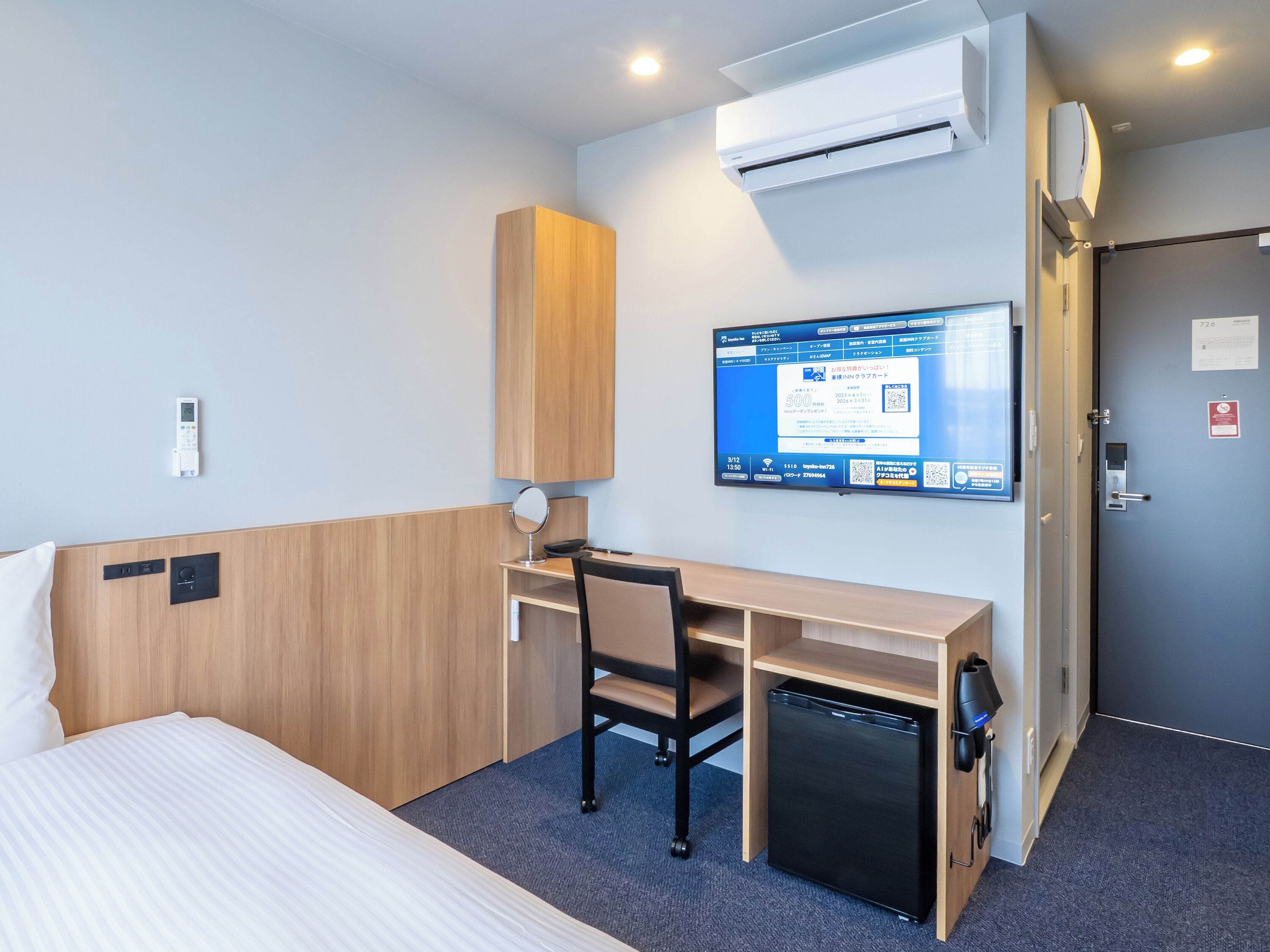 A hotel room with a bed, a desk with a chair, a small refrigerator, an air conditioning unit, a television, and a door with a lock.