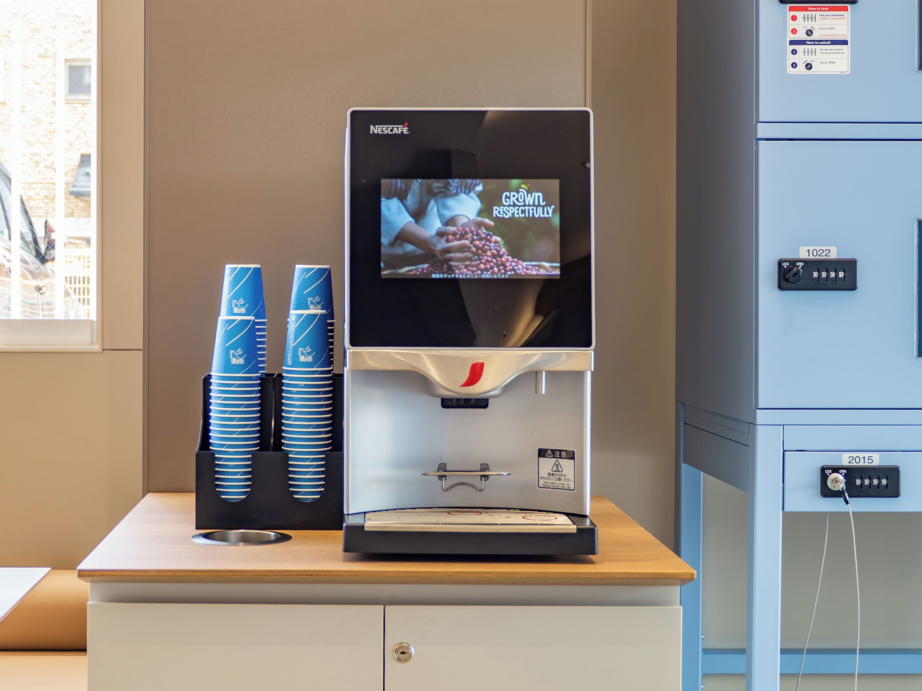 Una máquina de café Nescafé con una pantalla que muestra 'GROWN RESPECTFULLY', tazas azules apiladas y un panel de control con un interruptor de bloqueo.