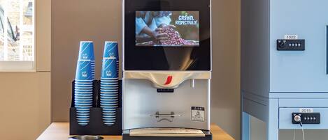 Una máquina de café Nescafé con una pantalla que muestra 'GROWN RESPECTFULLY', tazas azules apiladas y un panel de control con un interruptor de bloqueo.