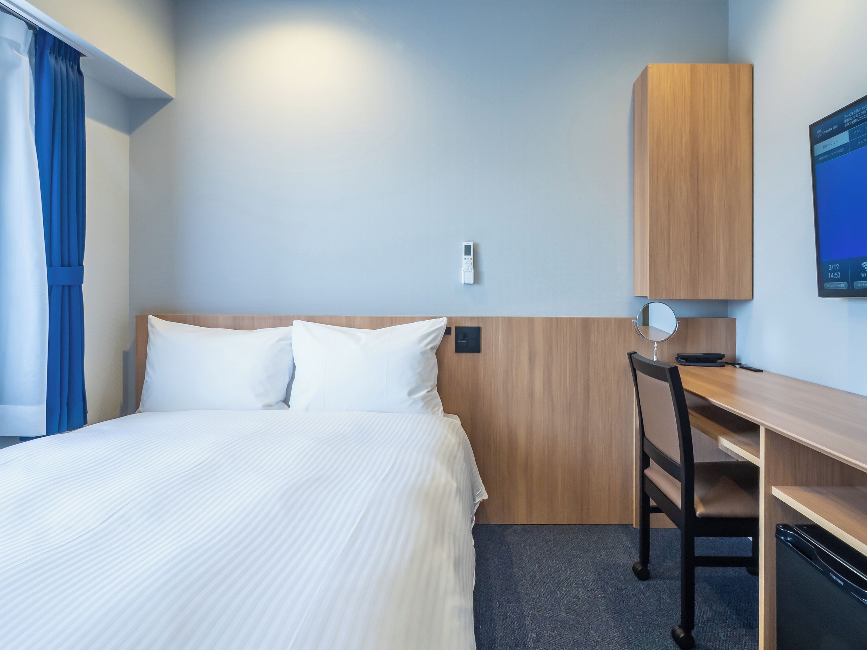 Habitación de hotel con cama, escritorio, silla, televisor montado en la pared y termostato.