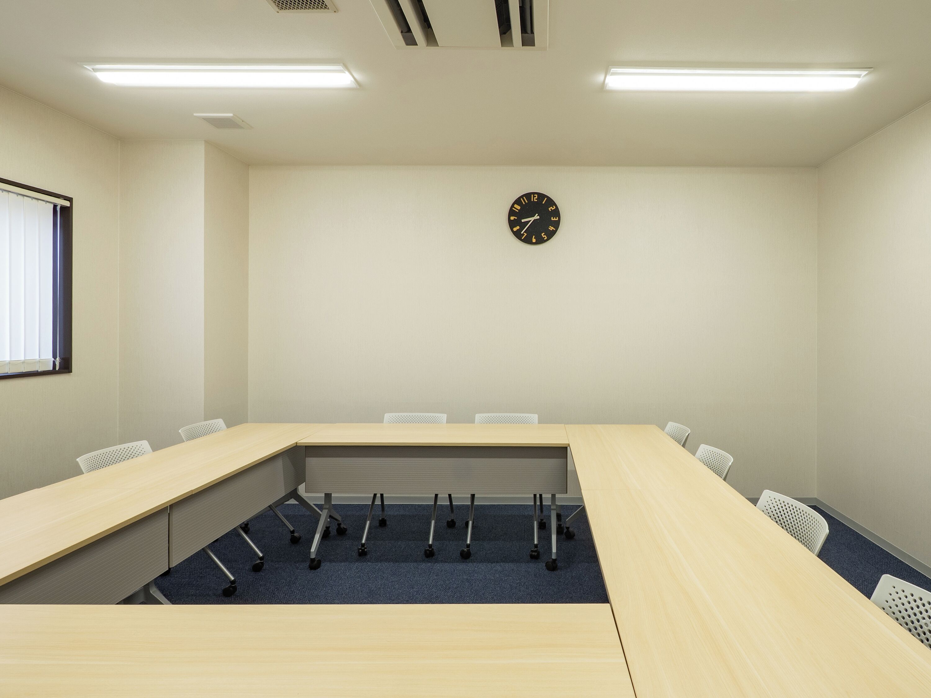 A conference room with a long table, chairs, a clock, and a window with blinds.