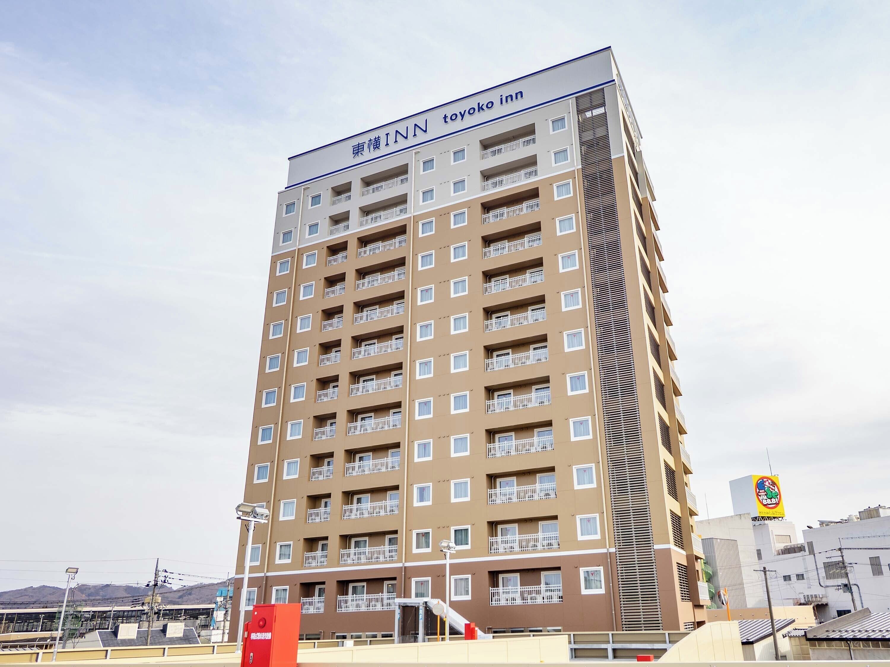 A tall hotel building with multiple balconies, a sign reading 'Toyoko Inn,' and a view of the surrounding urban area.
