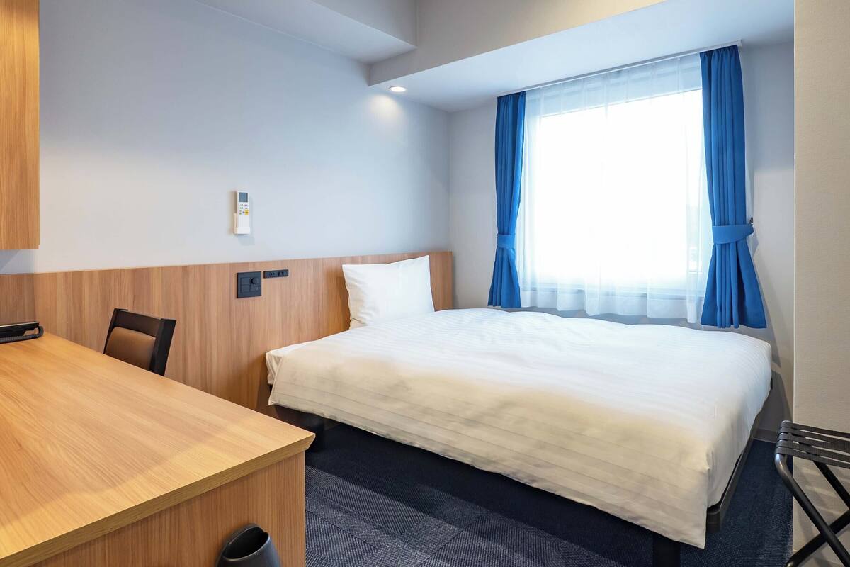 A hotel room with a bed, a desk, a chair, a window with curtains, and a control panel on the wall.