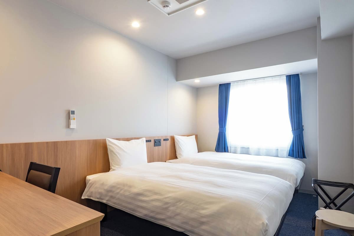 A hotel room with two beds, a desk, a chair, a window with curtains, and a ceiling light.