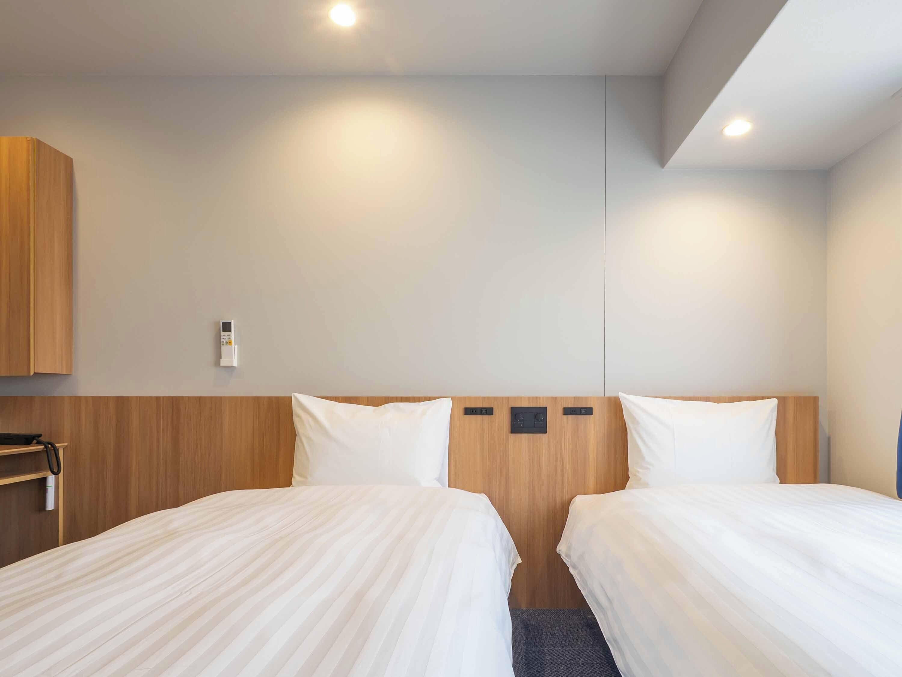 A hotel room with two beds, a wooden headboard, a wall-mounted thermostat, and a small shelf with a bottle.