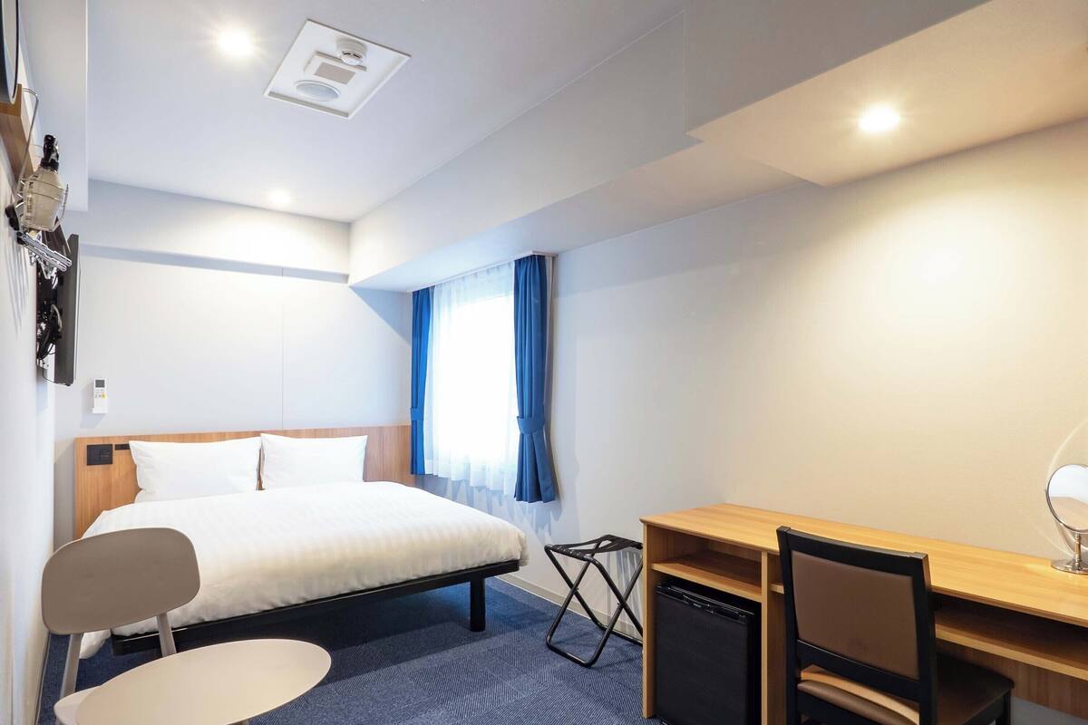 A hotel room with a bed, a desk, two chairs, a small table, a compact refrigerator, and a ceiling light.